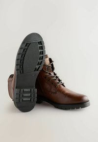 Brązowe skórzane buty na sznurówki z okrągłym czubkiem, fakturowaną czarną gumową podeszwą i widocznymi detalami przeszycia. Podeszwa ma wzór bieżnika.