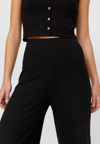 Nära bild av en person som bär svarta ribbade högmidjade byxor och en kort, knäppt topp som visar magen och armarna.