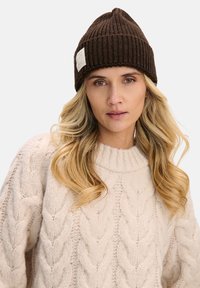 Brązowa dzianinowa czapka z prążkowaną fakturą i małą metką z przodu, noszona na jasnym, grubym swetrze z warkoczowym splotem.