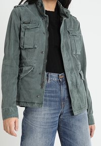 Groene canvas jas met meerdere frontzakken, een ritsluiting en een staande kraag, gedragen over een zwarte trui en blauwe jeans.