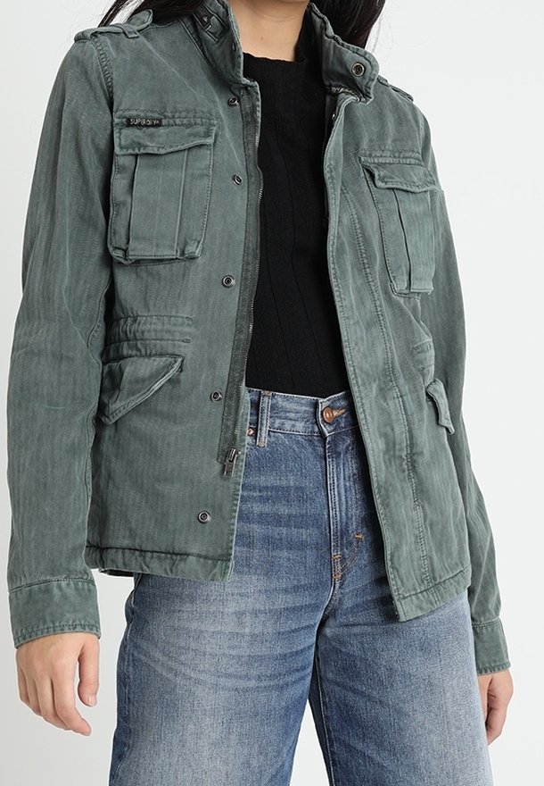 Groene canvas jas met meerdere frontzakken, een ritsluiting en een staande kraag, gedragen over een zwarte trui en blauwe jeans.