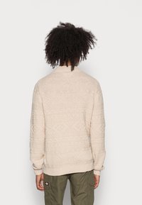 Beige stickad tröja med en strukturerad mönster, ribbade muddar och nederkant, samt standard passform. Baksidan visar upp intrikata designdetaljer.