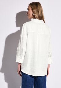 Weißes Leinenhemd mit einem Rückenfalten, langen Ärmeln, die an den Manschetten aufgerollt sind, und einem abgerundeten Saum. Dazu werden tiefblaue Jeans getragen.