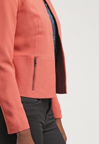 Veste ajustée de couleur corail avec un tissu texturé, dotée d'une poche zippée et d'un design court. Accompagnée d'un haut foncé et d'un pantalon noir.