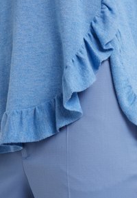 Haut tricoté bleu clair avec ourlet volanté sur un côté, associé à un pantalon texturé bleu clair. Les deux tissus semblent doux et légers.