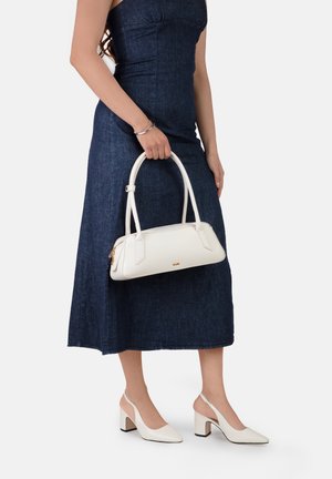 Žena u tamnoj traperskoj haljini bez rukava drži bijelu kožnu torbu sa zlatnim patentnim zatvaračem i nosi bijele slingback cipele s blok petom.