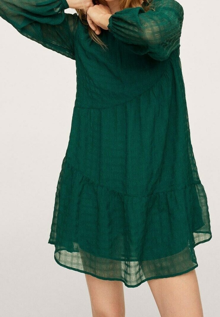 Femme portant une robe longue manches, verte foncée, transparente, avec une jupe à volants, debout, les bras levés près du visage.