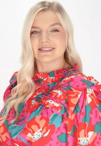 Textil blúz színes virágmintával, vörös és zöld árnyalatokban, fodros nyakkivágással és rakott részletekkel. Simogató, könnyű anyag.