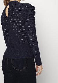 Námořnická modrá blůza s dlouhým rukávem a vycpanými rameny, s klíčovou dírkou na zádech a složitým vzorem řezaných diamantových tvarů.