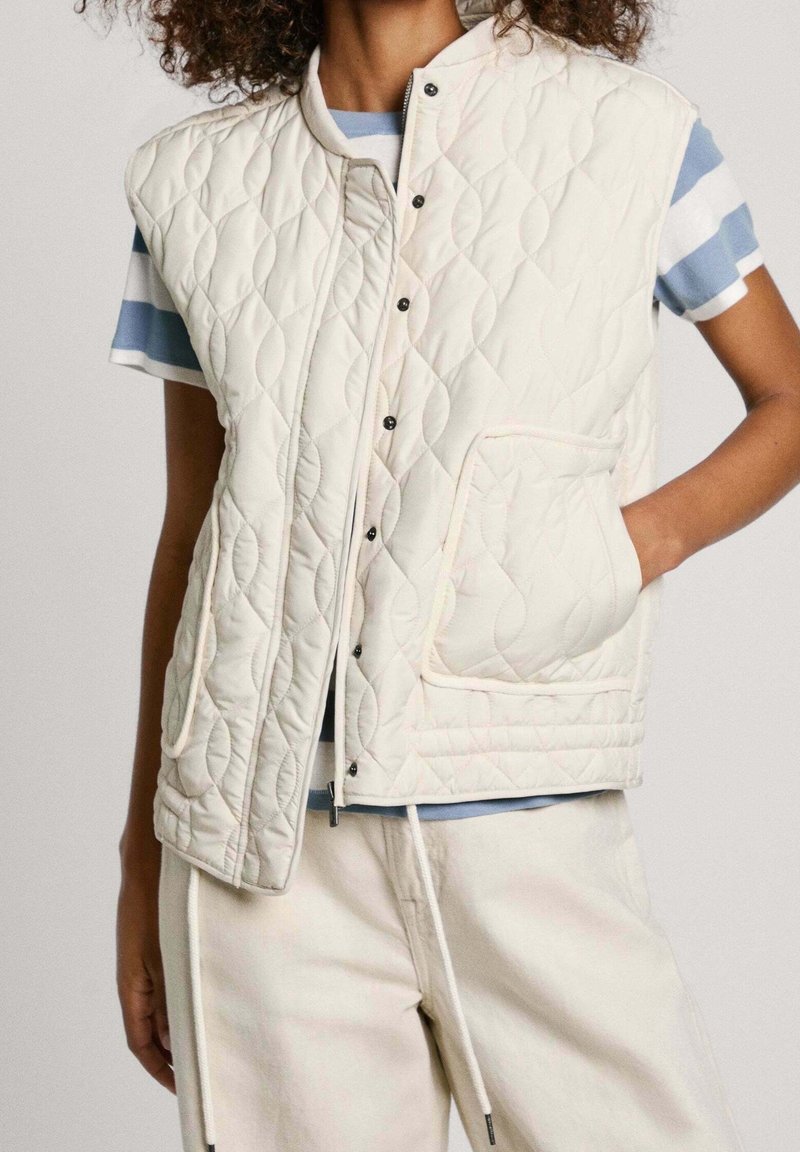Prošívaná bezrukávová vesta v krémové barvě s stojáčkem, zdobená černými patentkami a přední kapsou. Nositá přes modrou a bílou pruhovanou košili.