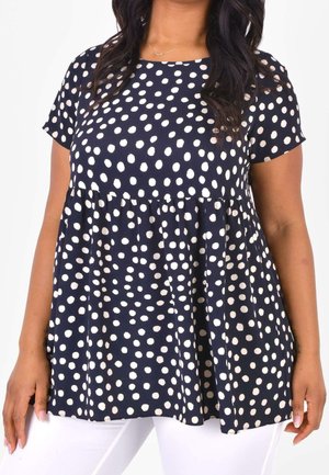 Donna che indossa un top blu navy a maniche corte con pois bianchi e pantaloni bianchi, mostrando la parte superiore del corpo e la zona centrale.