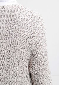 Ljust beige öppenstickad tröja med ett texturerat mönster, vida ärmar och en lätt drapering, visad från bakvyn.