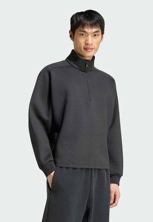 Joven con sudadera de cuello alto con cremallera en gris oscuro y pantalones oscuros a juego, de pie con una mano en el bolsillo.