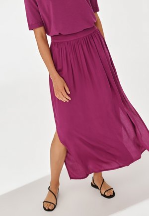 Femme portant une jupe midi fluide magenta et un haut assorti avec des sandales plates noires à brides, debout devant un fond blanc uni.