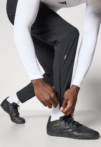 Schwarze Sporthosen mit tailliertem Design, kombiniert mit schwarzen Fußballschuhen mit strukturierten Sohlen und weißen Socken, vor einem neutralen Hintergrund.