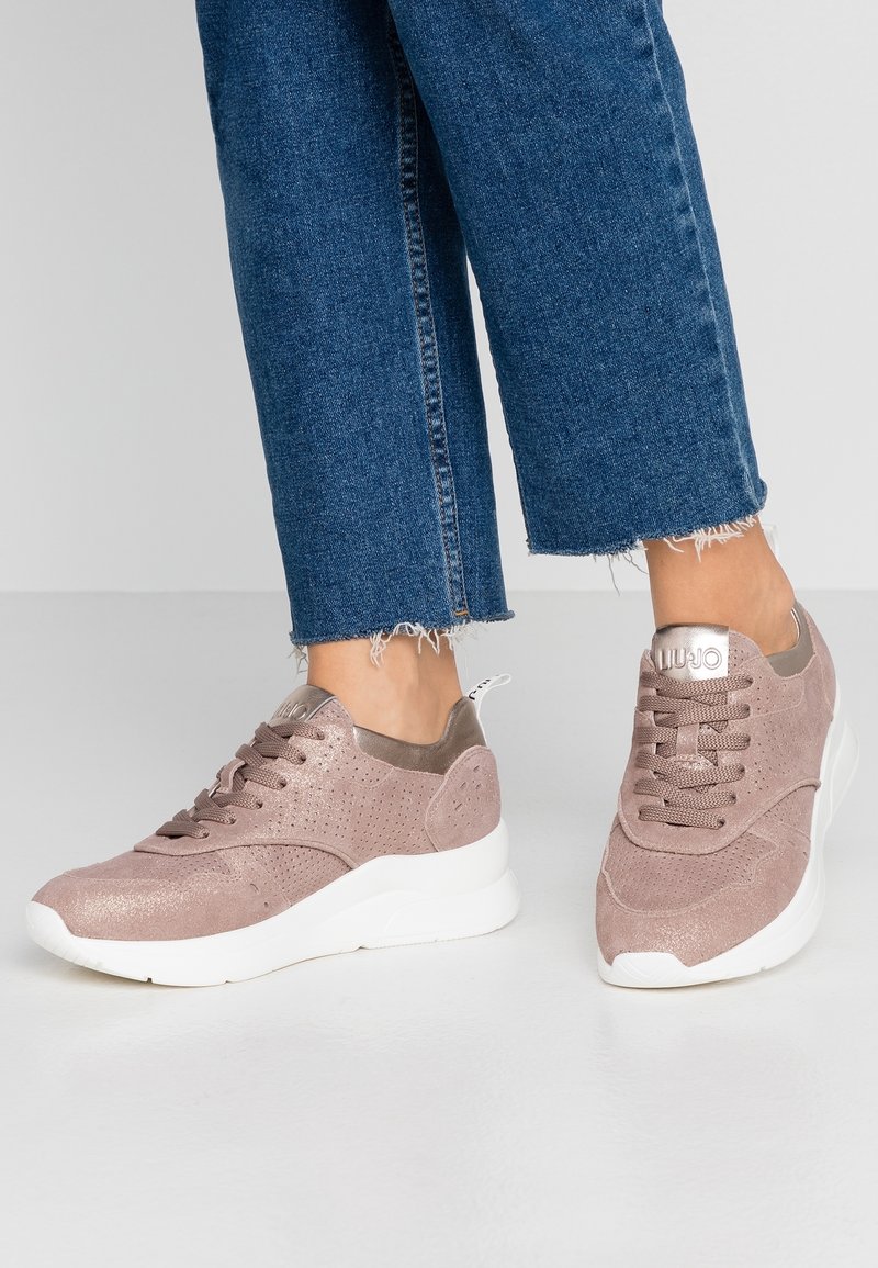 Femme portant un jean court bleu et des baskets en suède rose poudré avec des semelles blanches, debout sur un sol gris.