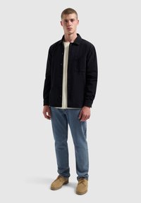 Zwarte corduroy button-up shirt met een borstzak, gedragen over een crèmekleurig T-shirt, gecombineerd met lichtblauwe jeans en beige schoenen.