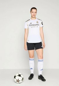 Bijela nogometna majica s crnim i zlatnim naglascima, crne kratke hlače i čarape s plavim prugama. Crne kopačke i lopta za nogomet crno-bijele boje u blizini.