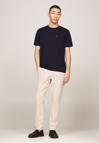 Tengerész kék pamut póló rövid ujjal, mellzsebkendővel, világos bézs slim-fit nadrággal és fekete slip-on cipővel párosítva. Egyszerű dizájn, minta nélkül.