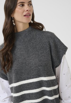 Pikkade pruunide juustega naine, kellel on seljas hõbedased ripatsiga kõrvarõngad, hallist koesilmaga ilma varrukateta topp valgete triipudega ja valge särk, millel on väiksed mustad mustrid.