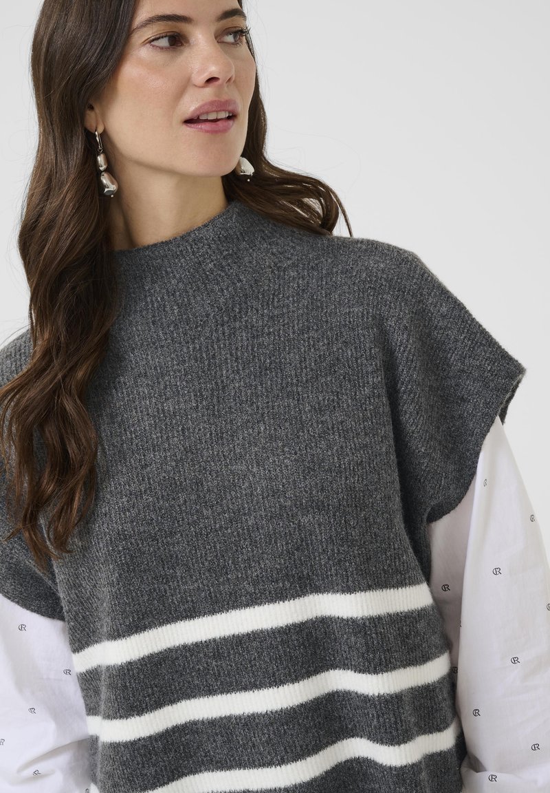 Sieviete ar gariem brūniem matiem, kas valkā sudraba karājas auskarus, pelēku adītu bezpiedurkņu topu ar baltām svītrām un baltu krekls ar maziem melniem rakstiem.