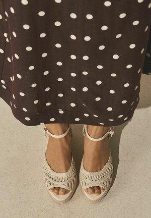 Füße, die cremefarbene gewebte offene Sandalen tragen, und ein brauner Rock mit weißen Polka-Punkten auf einem hell strukturierten Boden.