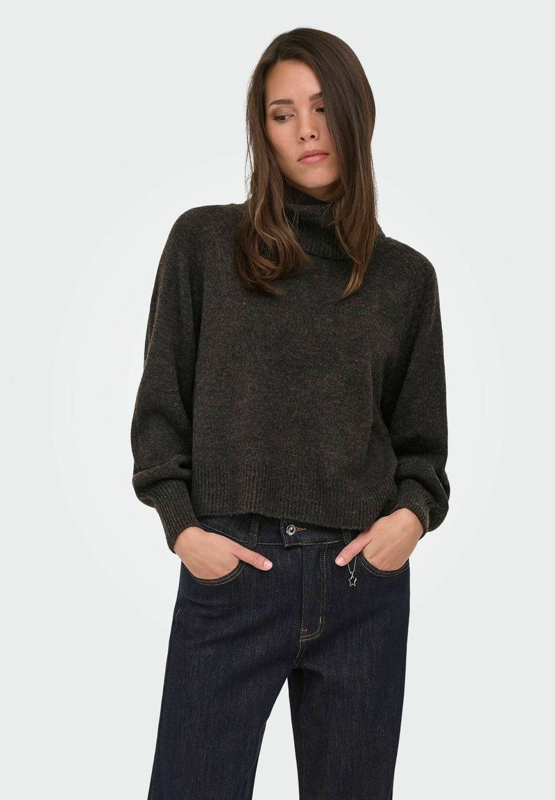 Femme portant un pull à col roulé gris foncé et un jean bleu foncé, les mains dans les poches, regardant légèrement vers le bas et sur le côté.