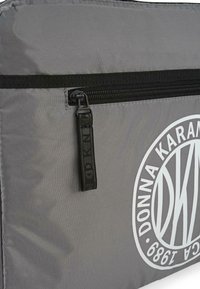 Bolsa de nailon gris con cremallera negra y tirador. Presenta un gran logo circular blanco con "Donna Karan" y "1998" impresos de manera prominente.
