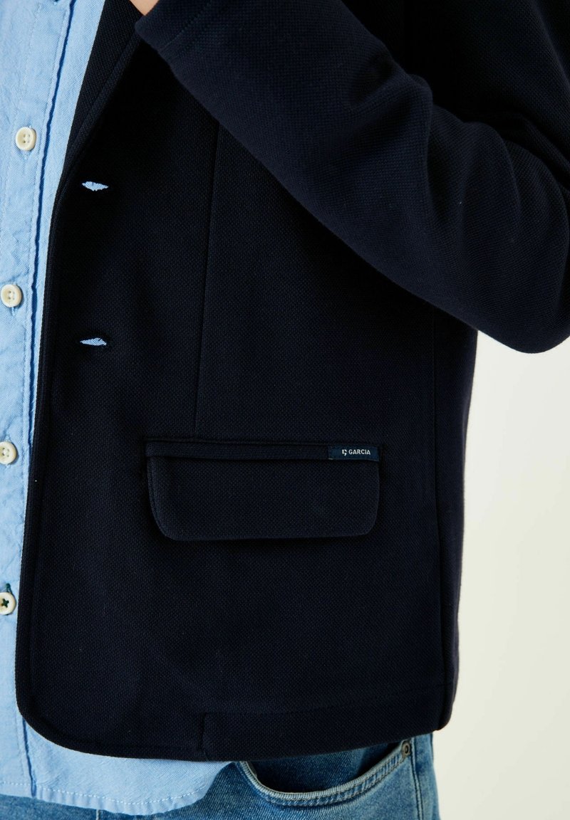 Giacca di colore blu navy con un taschino frontale, chiusura a bottoni e una camicia azzurra chiara sotto. L'etichetta con il logo sul taschino rivela i dettagli del marchio.