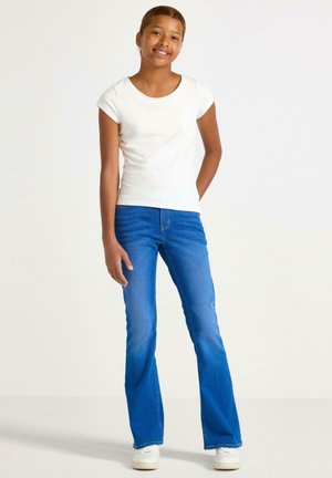 Smilende pige stående med den ene hånd bag ryggen, iført hvid kortærmet skjorte, blå vide jeans og hvide sneakers på ensfarvet baggrund.