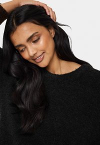 Crni plišani pulover s teksturiranim završetkom i okruglim izrezom. Duga, valovita tamna kosa slijeva se preko ramena. Glatka koža s suptilnim sjajem.