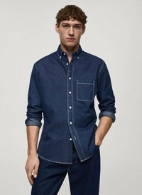 Temno modra srajca na gumb z dolgimi rokavi, spredaj žep na prsih, beli šivi in detajli s tremi zavoji. Obuta z usklajenimi jeans hlačami.