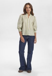 Blusa verde chiaro con stampa floreale, maniche a palloncino corte e scollatura a V, abbinata a jeans svasati blu scuro e scarpe marroni.