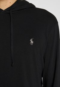 Svart hoodie i mjukt material med dragsko i huvan. En liten, silverfärgad broderad logga är placerad på vänster bröstomr åde.