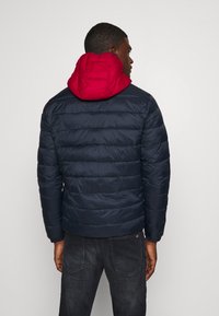 Tumes sinine puffer jakkk, millel on punane kapuuts, horisontaalselt tikitud sektsioonidega, sile tekstuur ja elastsed mansetid. Tagantvaade.