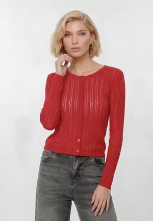 Blond žena v červenom pletenom kardigane na gombíky s dlhými rukávmi a vyblednutých čiernych džínsoch na jednofarebnom pozadí.