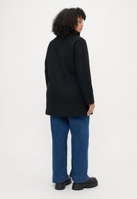 Cappotto di lana nero con un design strutturato, maniche lunghe e taglio dritto, abbinato a jeans in denim blu e scarpe con plateau nere e chunky.