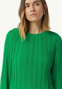 Robe plissée verte à manches longues et décolleté rond, avec des lignes verticales pour la texture et un tissu lisse et fluide.