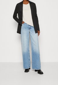 Czarna marynarka na jasnobeżowej koszulce z małym logo, zestawiona z szerokimi, jasnoniebieskimi jeansami i czarnymi botkami.