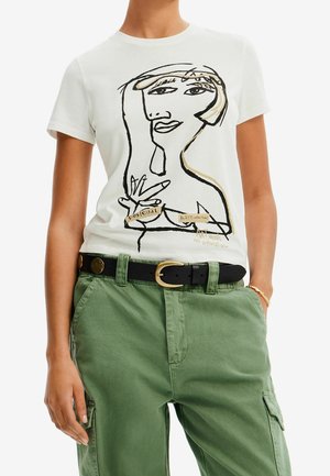 Vrouw draagt een wit T-shirt met een abstract lijntekening van een gezicht en tekst, gecombineerd met groene cargobroek en zwarte riem met gouden gesp.