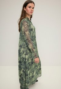 Vestido largo verde con una textura transparente, que presenta un patrón abstracto en tonos más oscuros y mangas largas. Suave caída en el dobladillo.