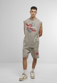 Beige sleeveless top with red and gray graphic, paired with matching beige shorts featuring a red design. Footwear: beige sneakers.