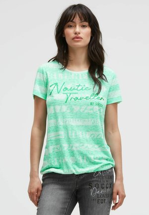 Woman with dark hair wearing a mint green striped t-shirt with "Nautic Traveller" text and gray jeans with printed text.