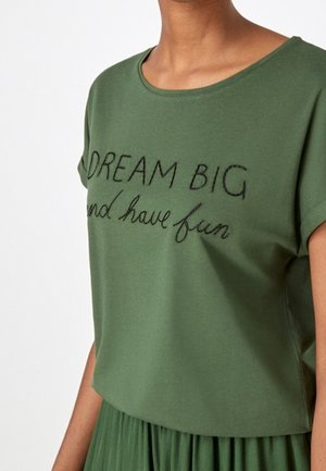 Haut vert à manches courtes avec texte brodé noir « DREAM BIG and have fun » porté par une personne à la peau de ton moyen, sur fond blanc.
