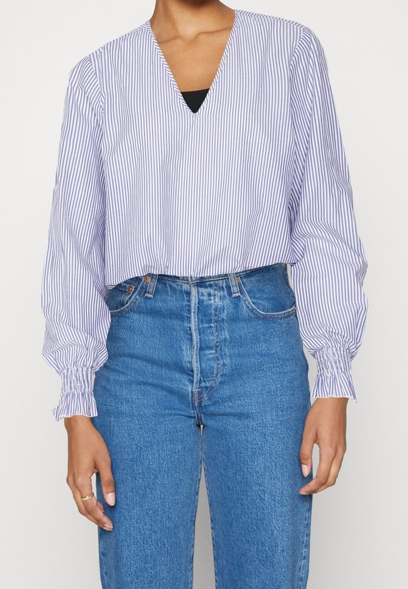 Femme portant un chemisier blanc à manches longues avec de fines rayures verticales bleues et un jean bleu taille haute, les mains détendues le long du corps.