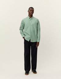 Zaļi un balti svītrots ilgs piedurkņu krekls ar pogājamu apkakli un krūšuniekšu, kopā ar brīviem melniem biksēm un melnām kurpēm.