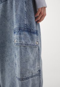 Šviesiai mėlynai deniminių salopetų su išblukusiu apdaila, su priekinėmis kišenėmis, sidabriniais detalėmis ir siuvinėtais akcentais.