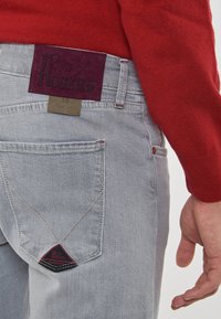 Nahaufnahme einer hellblauen Slim-Fit-Jeans mit Gesäßtasche und einem Roy Rogers-Lederpatch, neben der eine Person in einem roten Pullover steht.