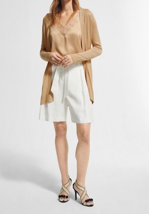 Cream silk camisole under a tan cardigan, paired with white pleated shorts and black strappy high-heeled shoes with pom-pom accents.