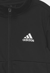 Zwarte ritsjack gemaakt van een gladde stof. Beschikt over een hoge kraag en een wit Adidas-logo op de linkerkant van de borst.